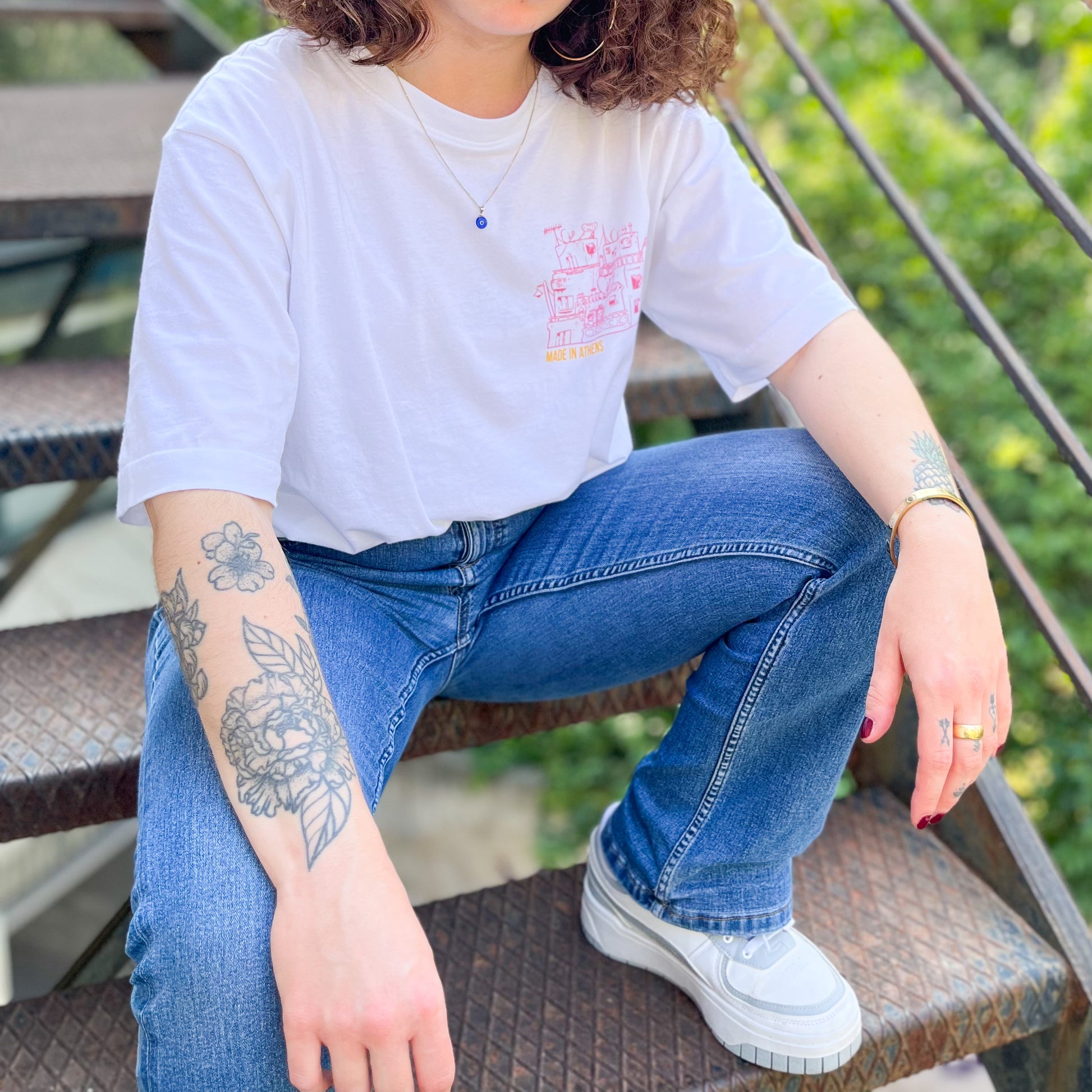 lady sitting with on stairs, the front design of the shirt is visible. Its is 3 small buildings of Athens only with a pink outline. Under it we can read "Made in Athens"