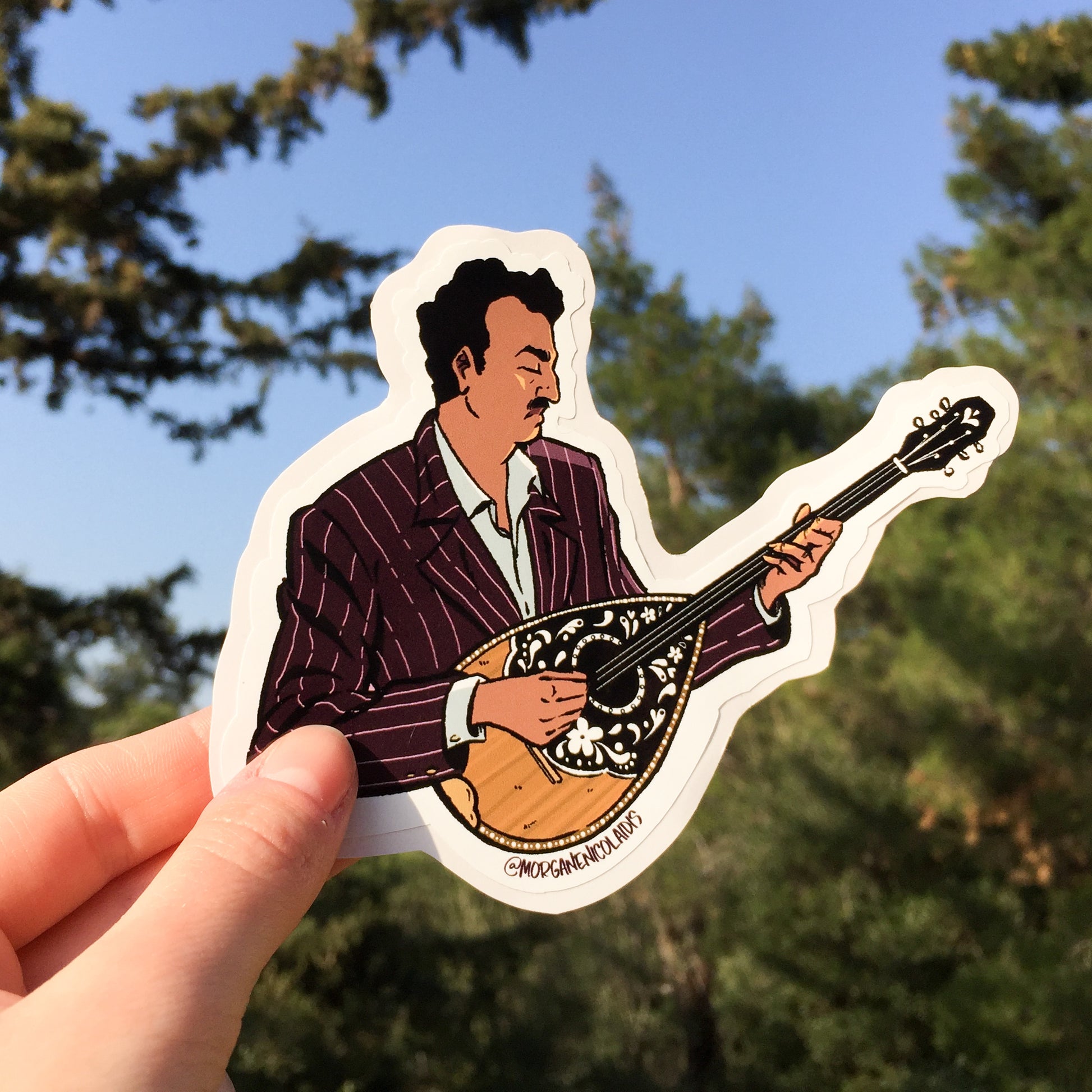 A hand holding the bouzouki player sticker. A lot of trees in the background.